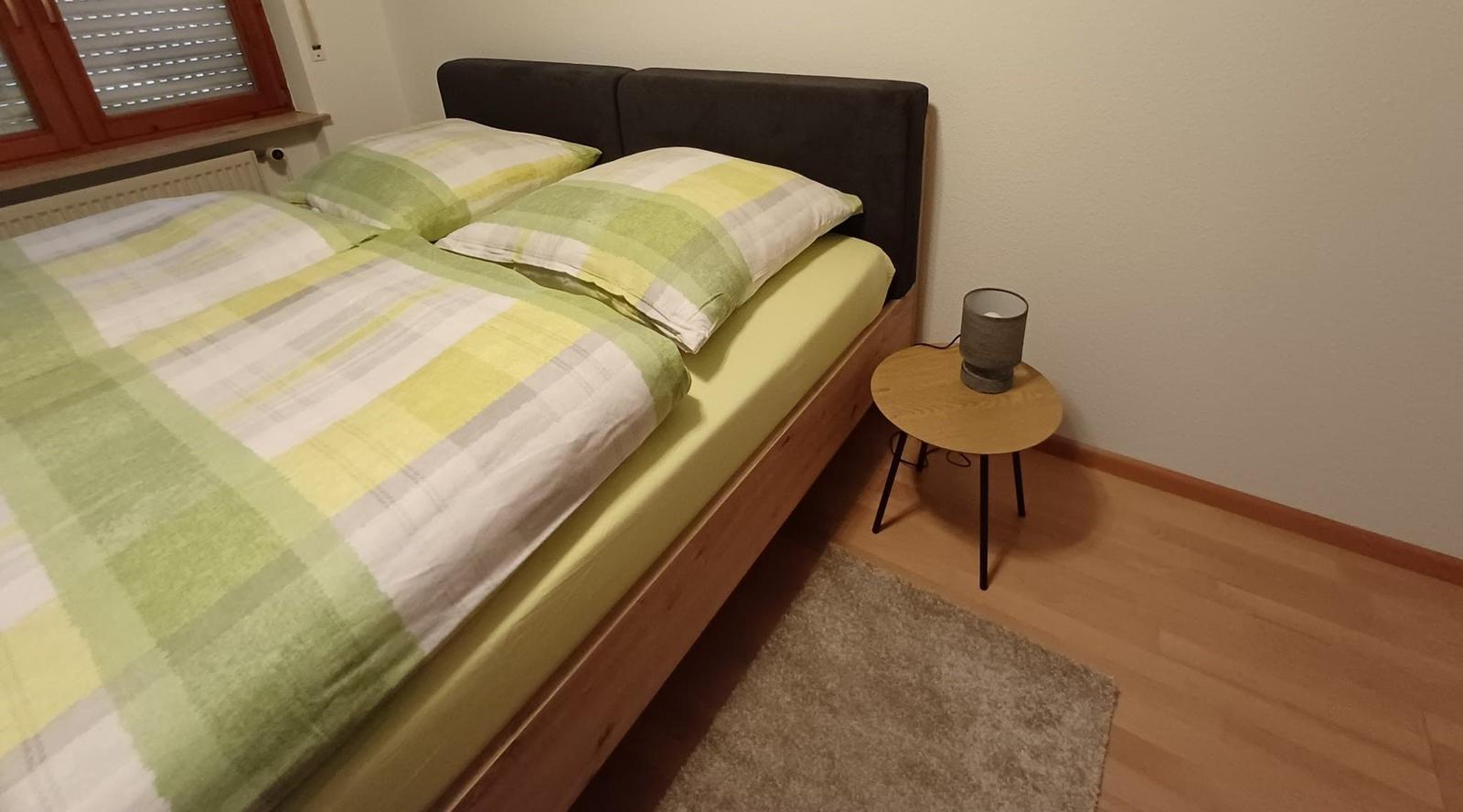 Ein Bett mit grün-weißem Bettzeug, Nachttisch und Fenster.