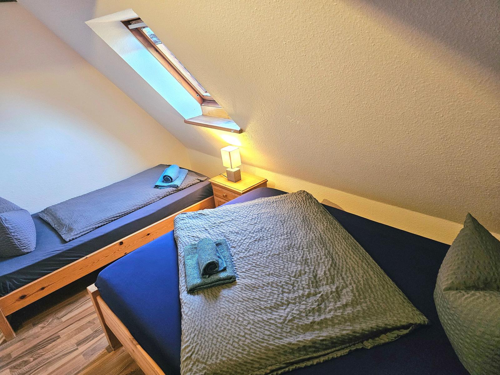 Zimmer mit zwei Betten, Dachfenster und Nachttisch mit Lampe.