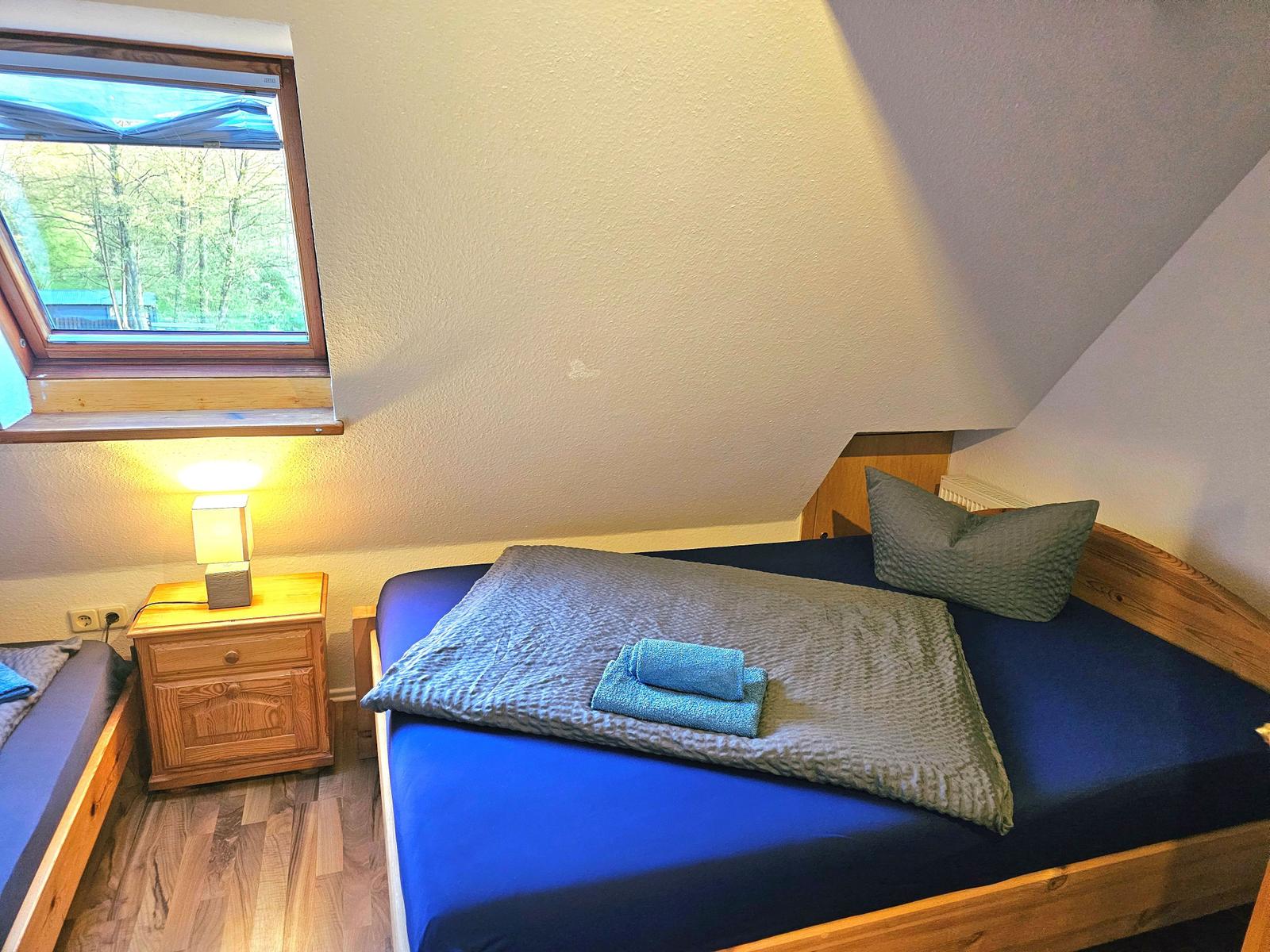 Schlafzimmer mit Dachschrägen, Bett, Nachttisch und Fenster mit Blick auf Wald.
