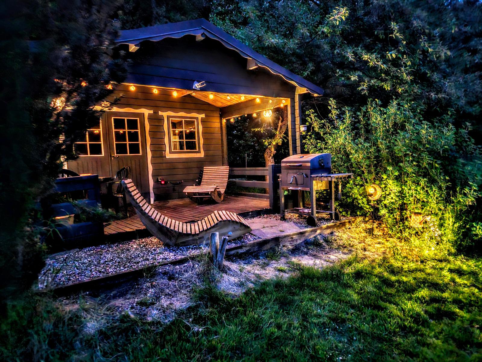 Hütte mit beleuchteter Veranda, Grill und Holzbank im Garten bei Nacht.