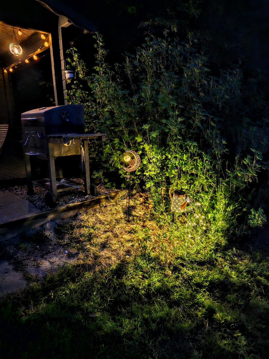 Gartenbereich bei Nacht mit Grill, Beleuchtung und Pflanzen.