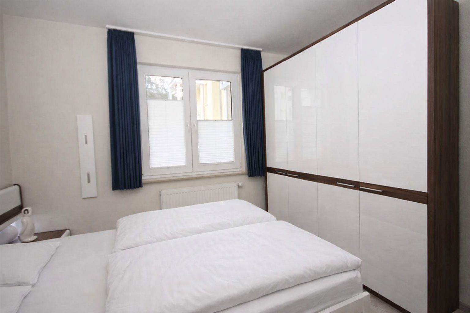 Schlafzimmer mit Doppelbett, weißen Bettwäsche, großen Schrank und Fenster mit blauen Vorhängen.