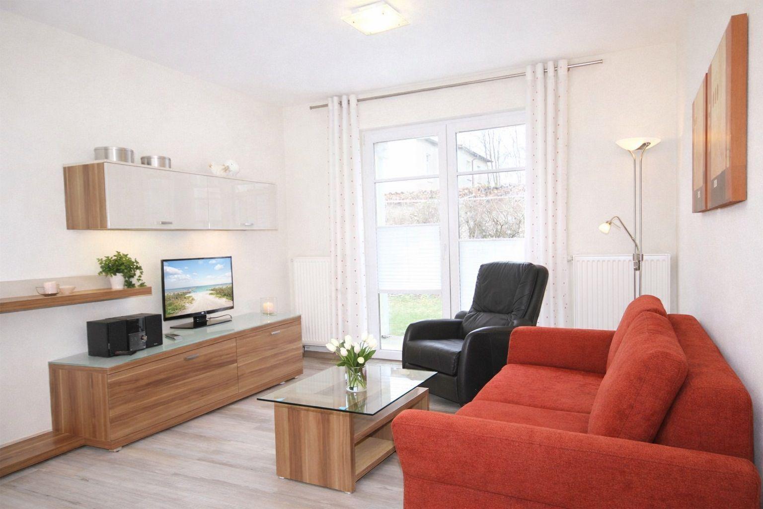 Wohnzimmer mit rotem Sofa, Glas-Tisch und Fernseher. Fenster mit weißen Vorhängen.