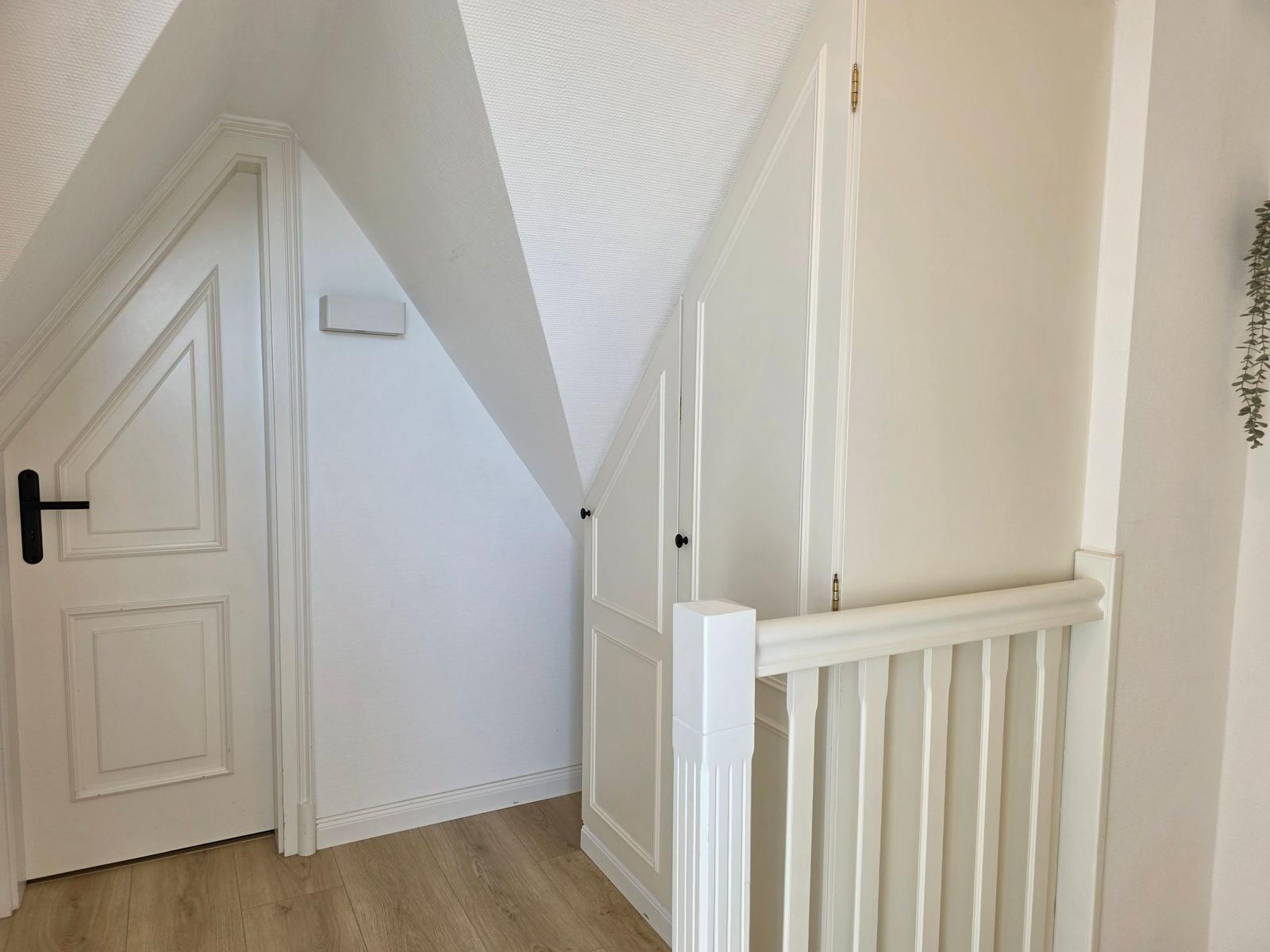 Treppenhaus mit weißen Türen und Geländer. Holzdielenboden und schräger Dachbereich.