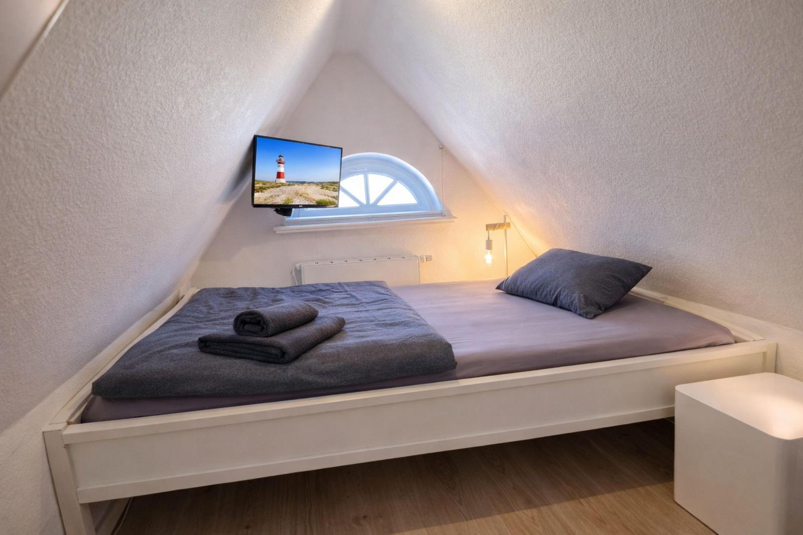 Schlafzimmer mit Dachschräge, Bett, Fernseher und Fenster.