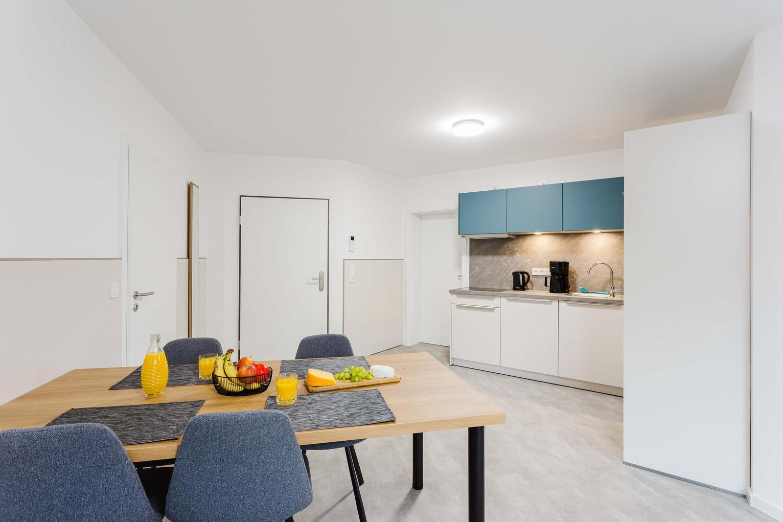 Essbereich mit Tisch, Stühlen und Obst. Küche mit blauen Schränken und Arbeitsplatte.