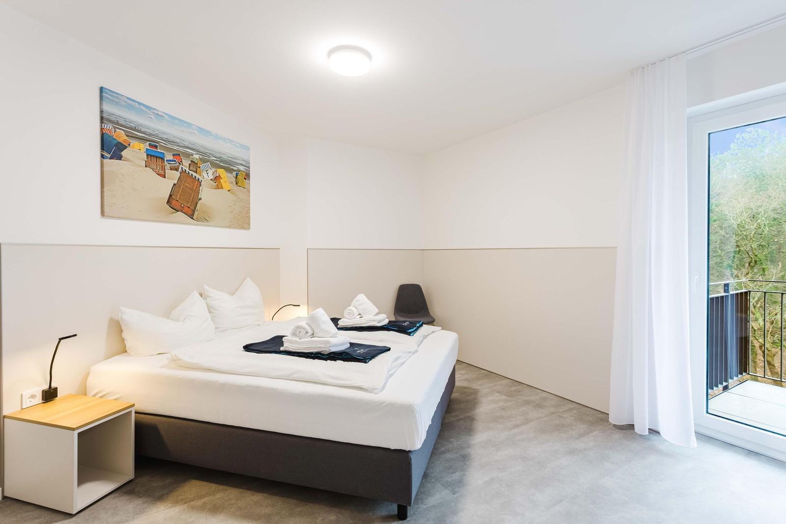 Schlafzimmer mit Bett, Nachttisch und Balkon. Wandbild zeigt Strandkörbe.