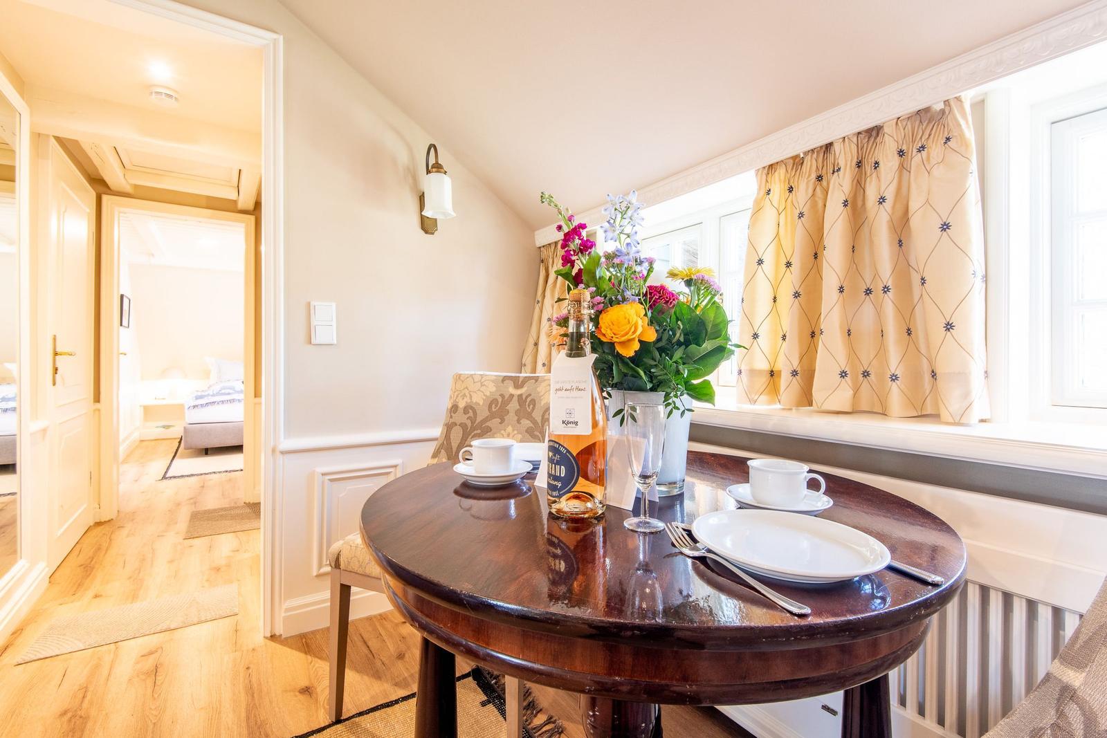 Rundtisch mit Tischdecke, Blumen und Getränken vor Fenster mit Vorhängen.