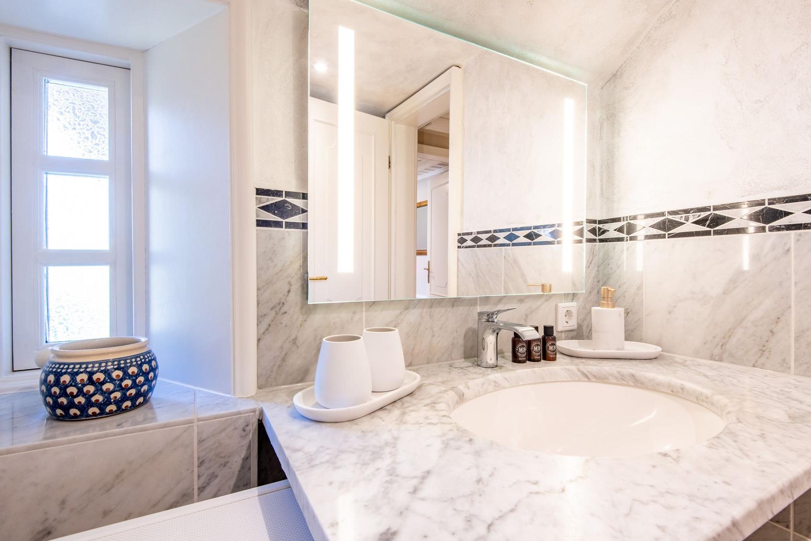 Badezimmer mit Marmorwaschtisch, beleuchtetem Spiegel und Fenster.