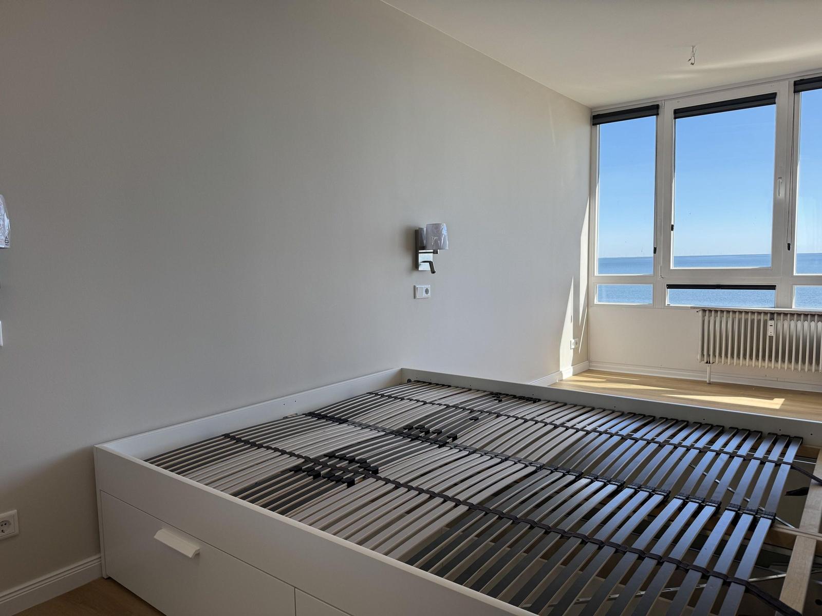 Bedroom with bed frame and sea view through large windows.