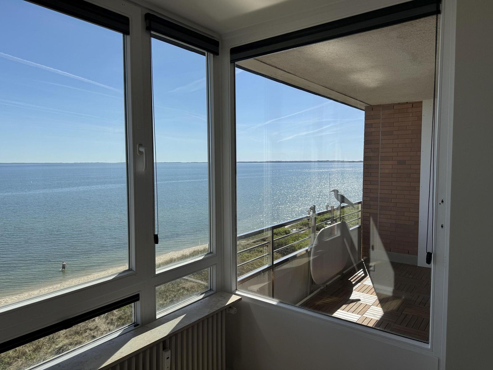 View of the sea through large windows and balcony with wooden deck.