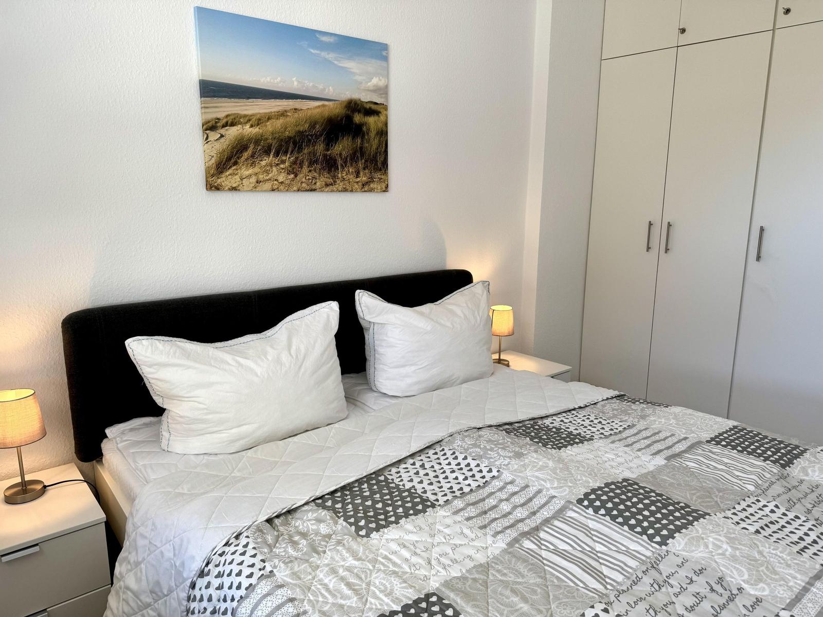 Schlafzimmer mit Bett, Nachttischen und Kleiderschrank. Wandbild zeigt Strand.