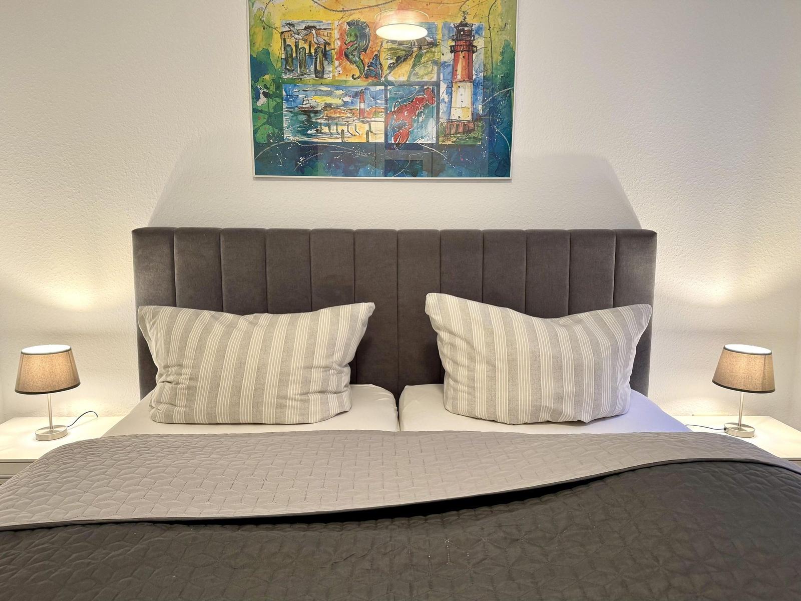 Doppelbett mit Kopfteil, Kissen und Decke. Wandbild mit Leuchte links und rechts.