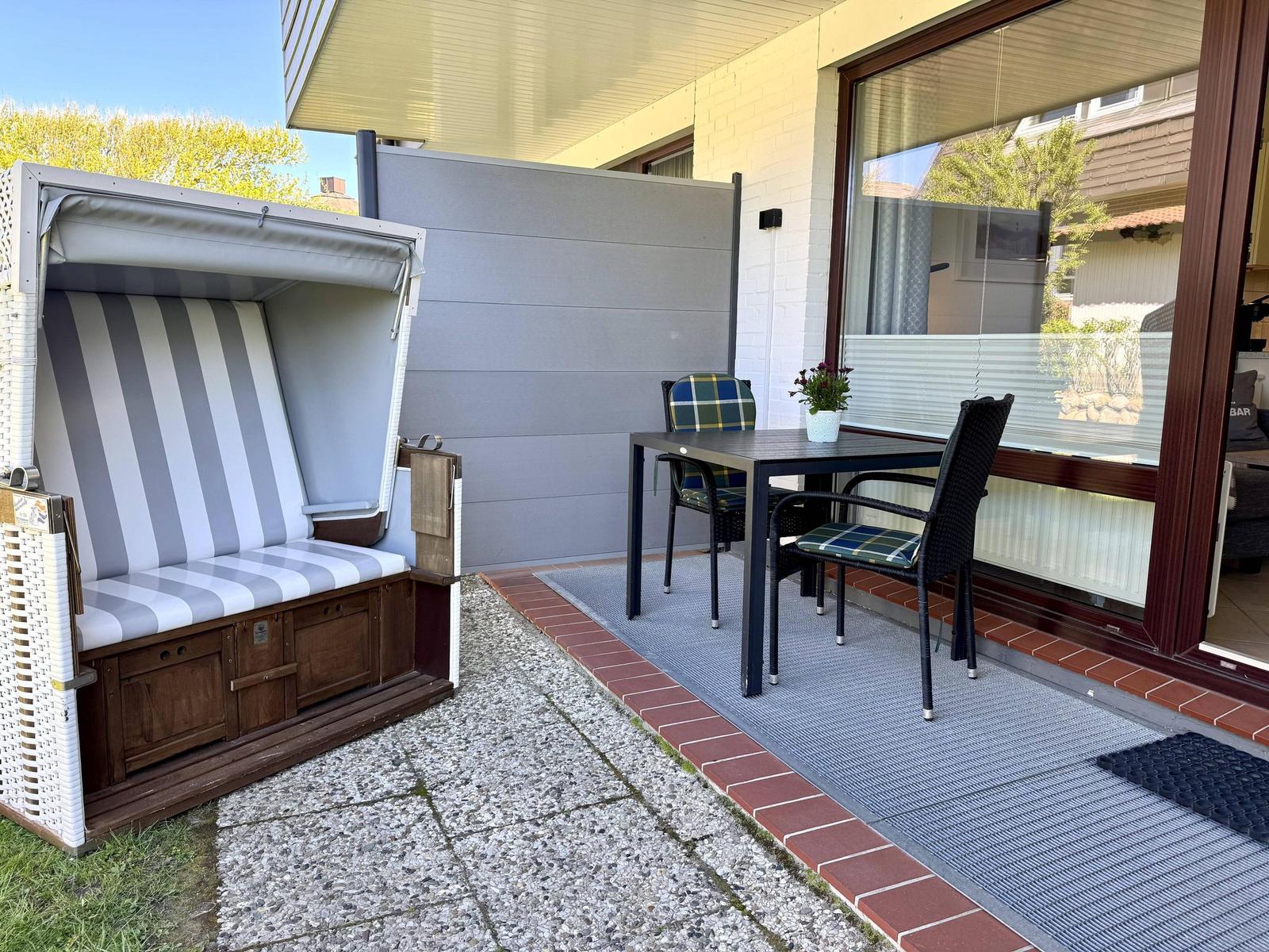 Terrasse mit Strandkorb, Tisch und zwei Stühlen. Bodenbelag aus Stein und Teppich.