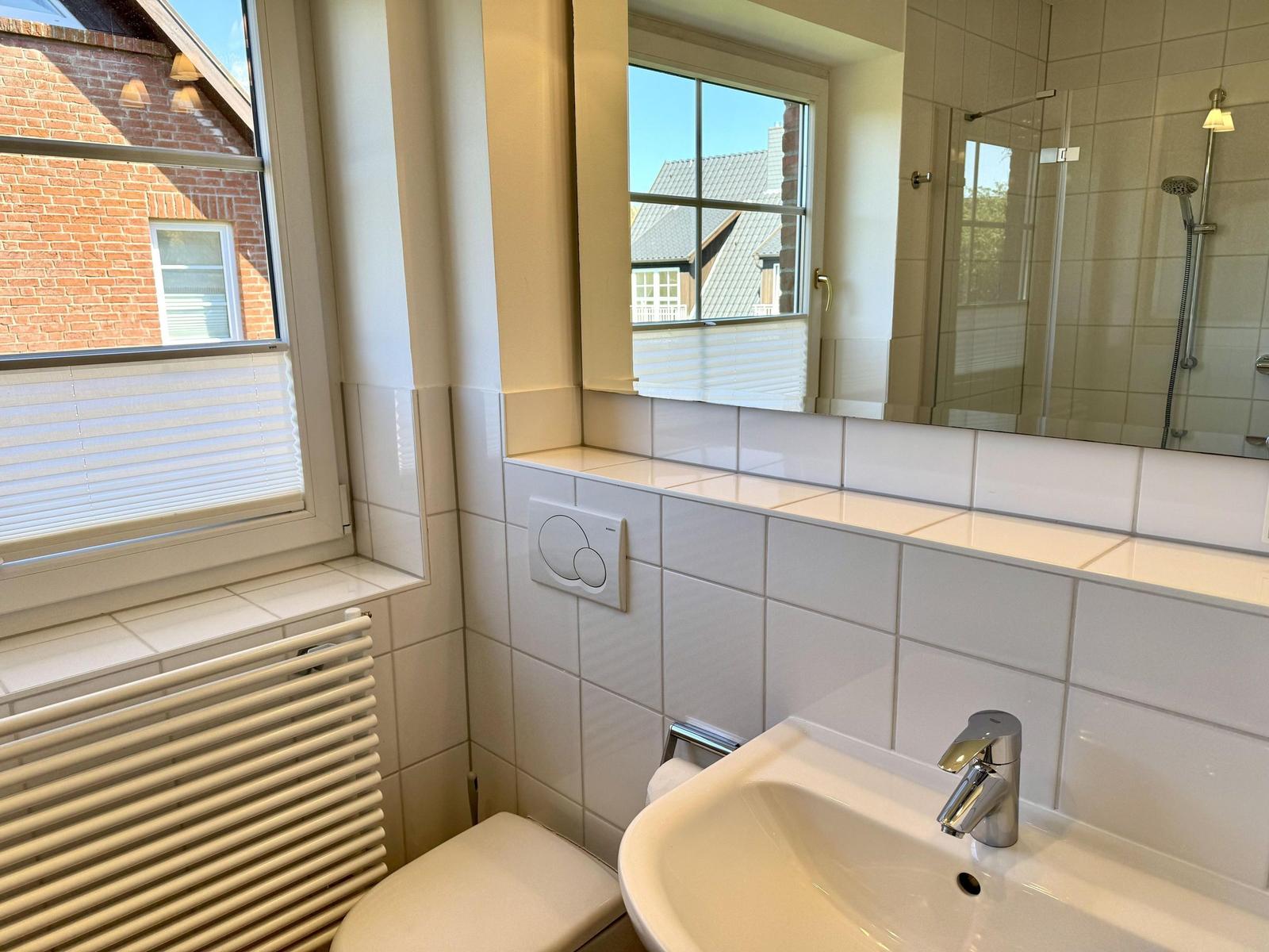 Badezimmer mit Waschbecken, Spiegel und Dusche. Fenster mit Jalousie.