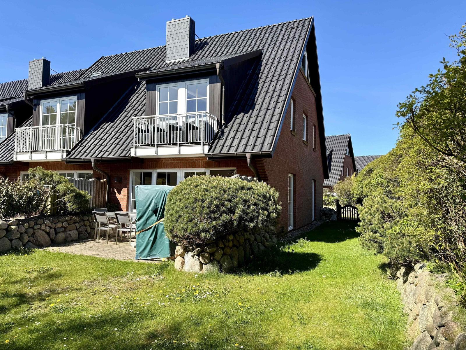 Haus mit Balkon, Garten und Steinmauer unter blauem Himmel.