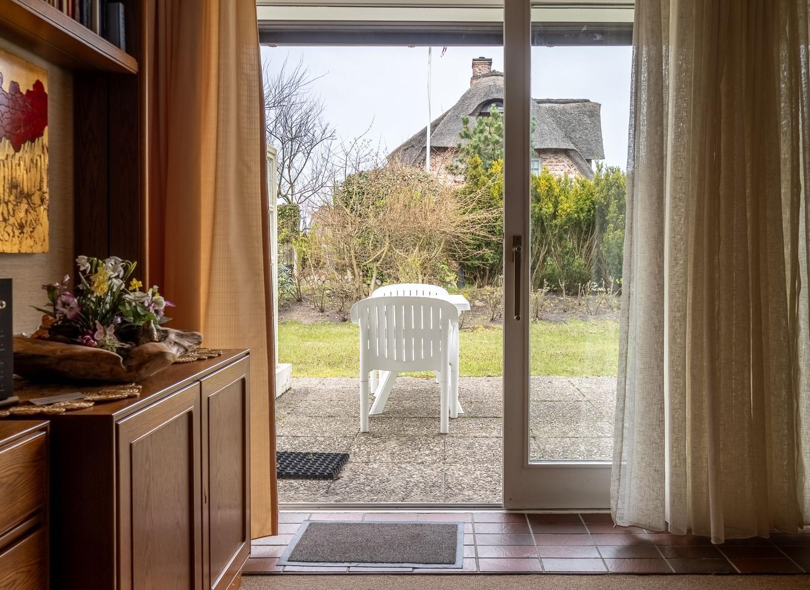 Durch Glastür zur Terrasse mit weißem Stuhl und Blick auf Gartenhaus.