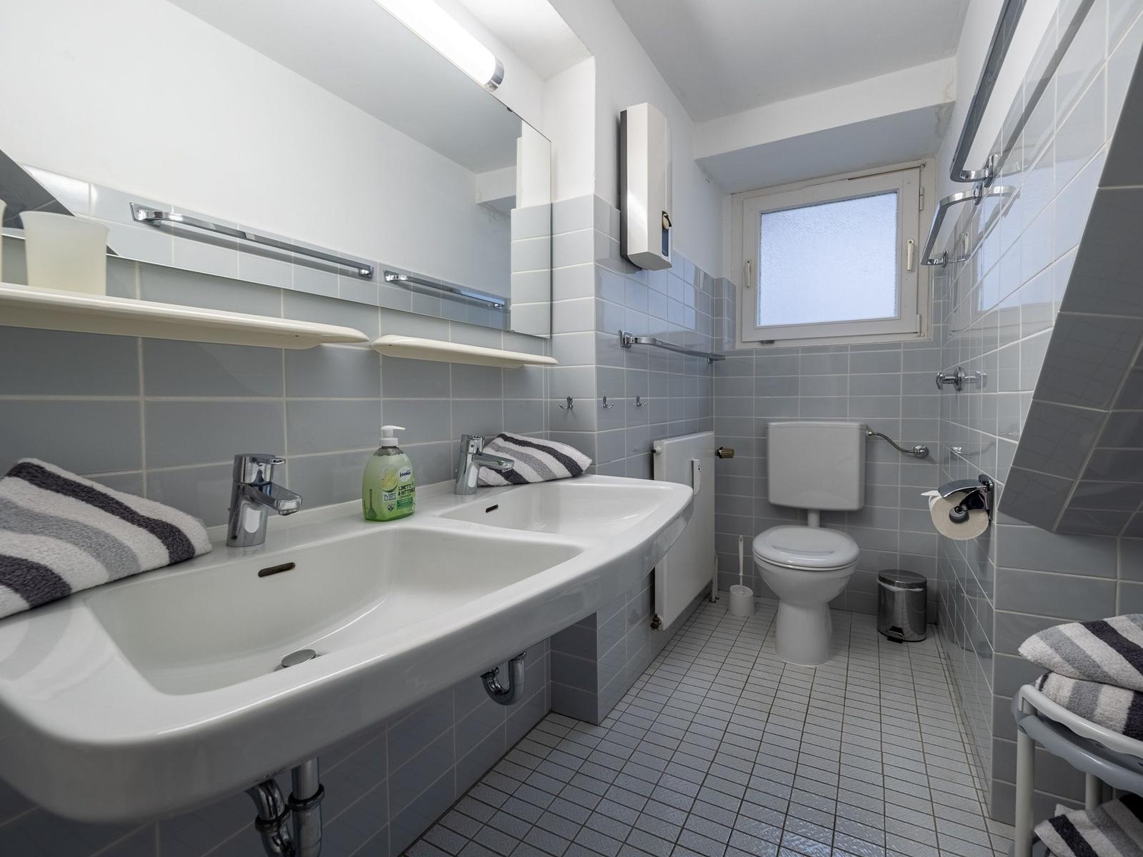 Badezimmer mit Waschbecken, Toilette und Dusche. Fliesenwand und Fenster sichtbar.