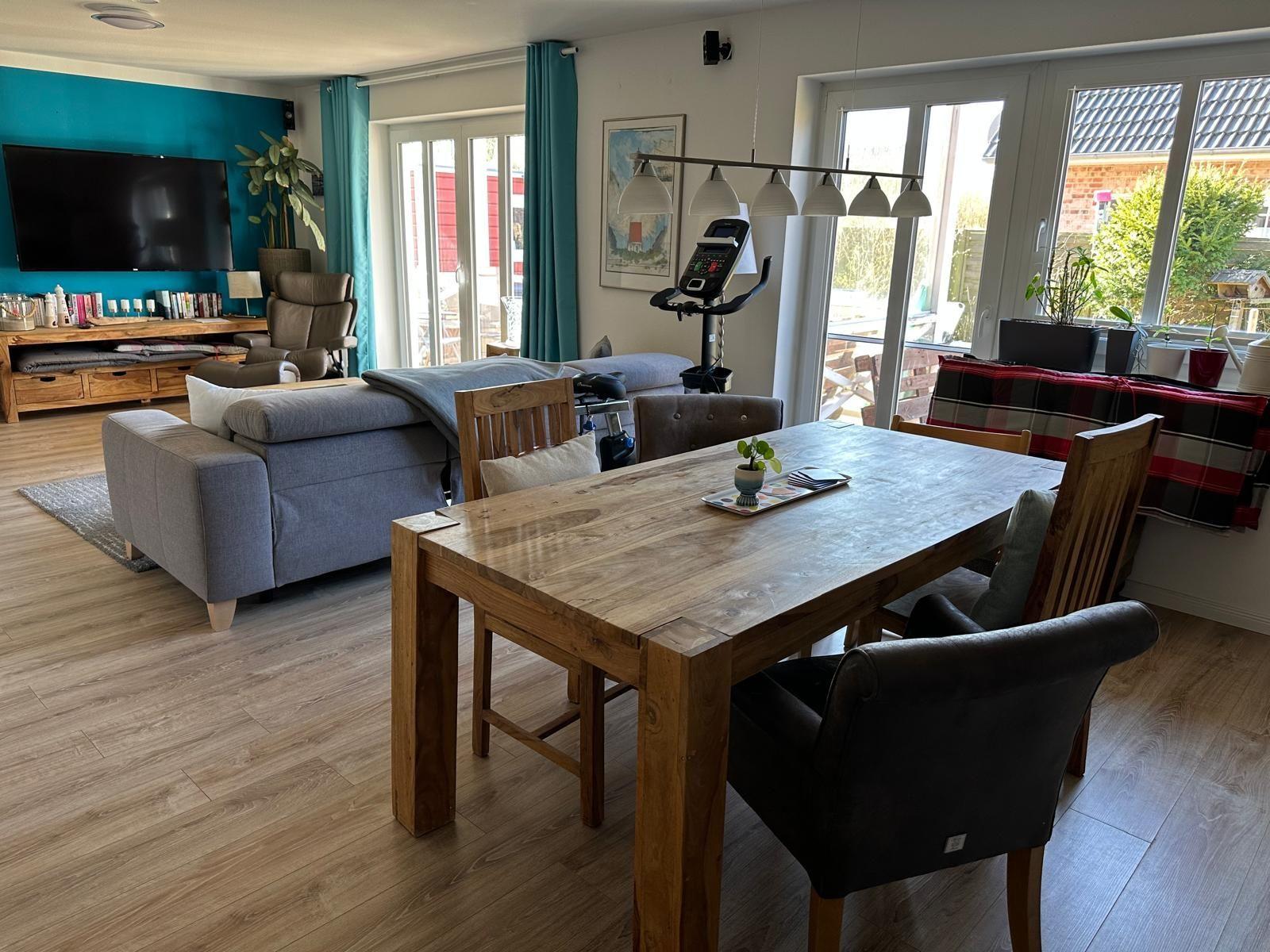 Essbereich mit Holztisch und Stühlen. Im Hintergrund Wohnbereich mit Sofa und TV.