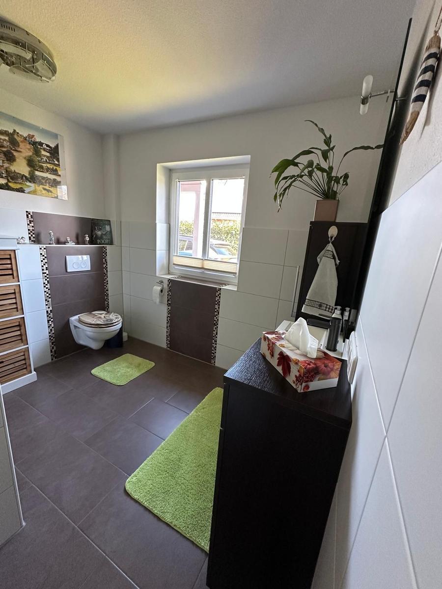 Badezimmer mit Toilette, Waschbecken und Fenster. Grün Teppiche und Pflanze.