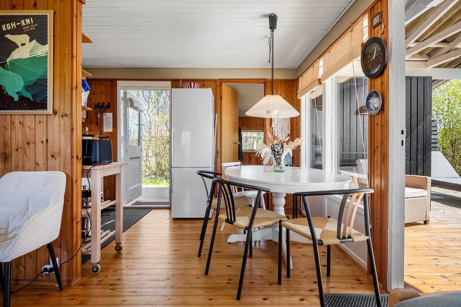 Dining area with round table, chairs, and refrigerator. Wooden walls and floor. Windows to terrace.