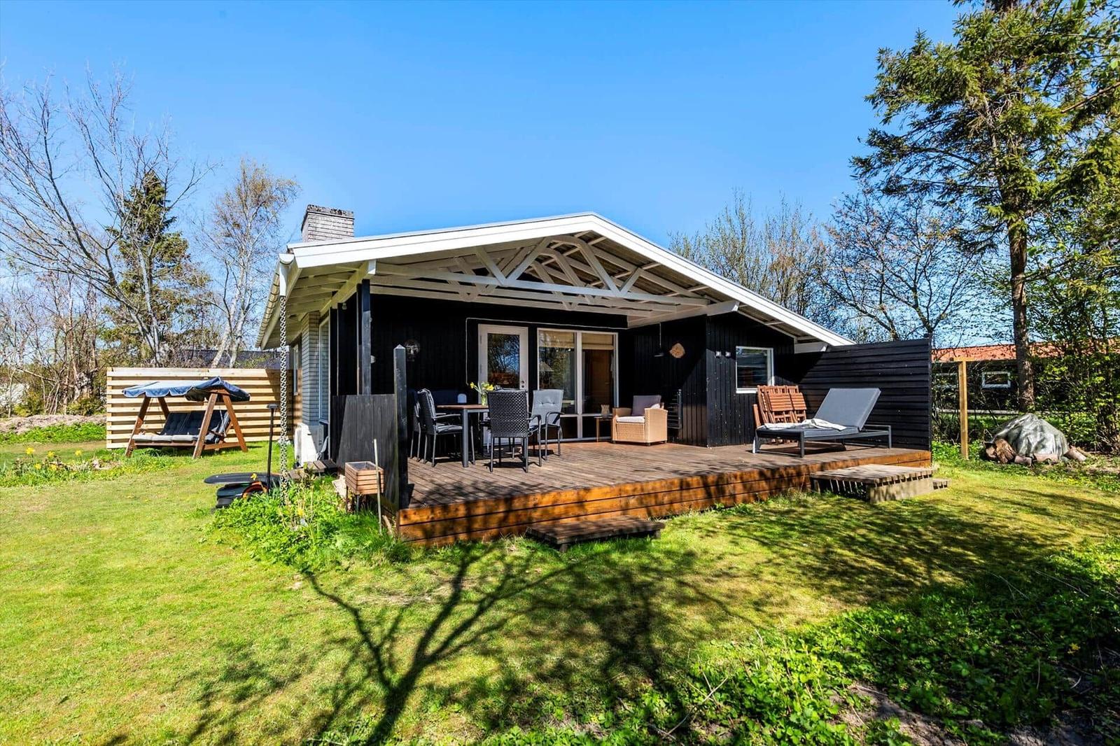 Modern cabin with wooden terrace, garden, and outdoor seating under blue sky.