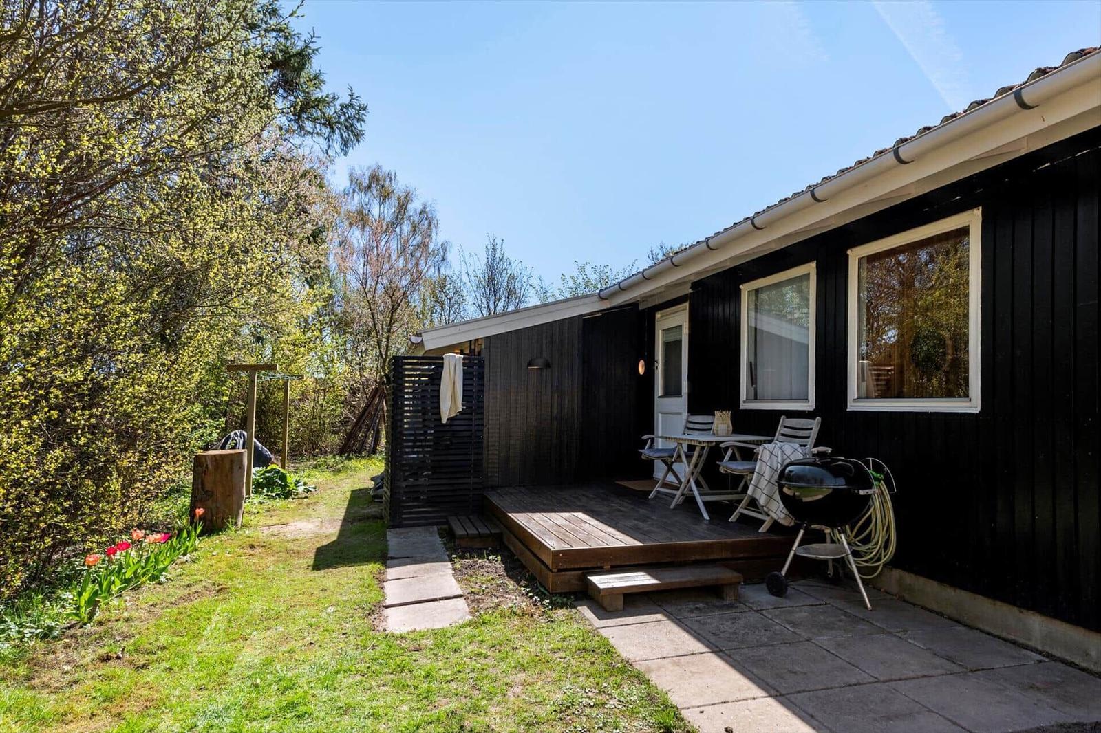 Black wooden cabin with terrace, grill, and garden. Table and chairs are ready.