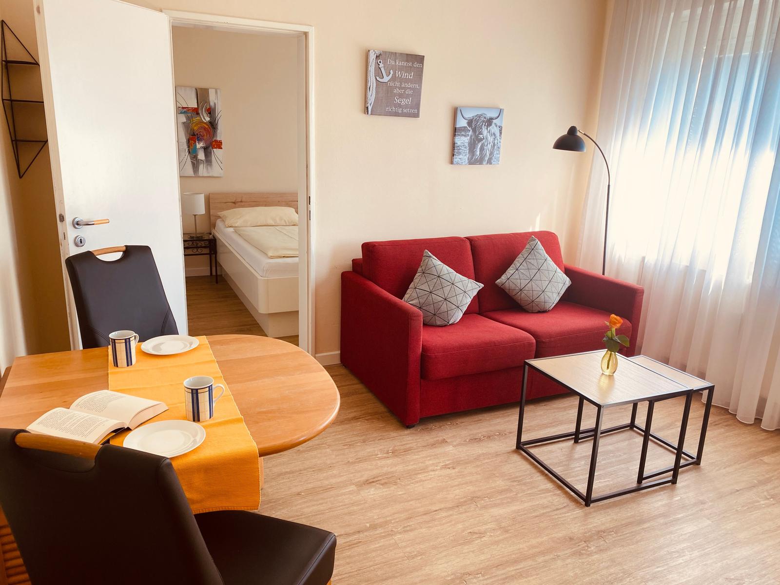 Wohnbereich mit rotem Sofa, Esstisch und Blick in Schlafzimmer.