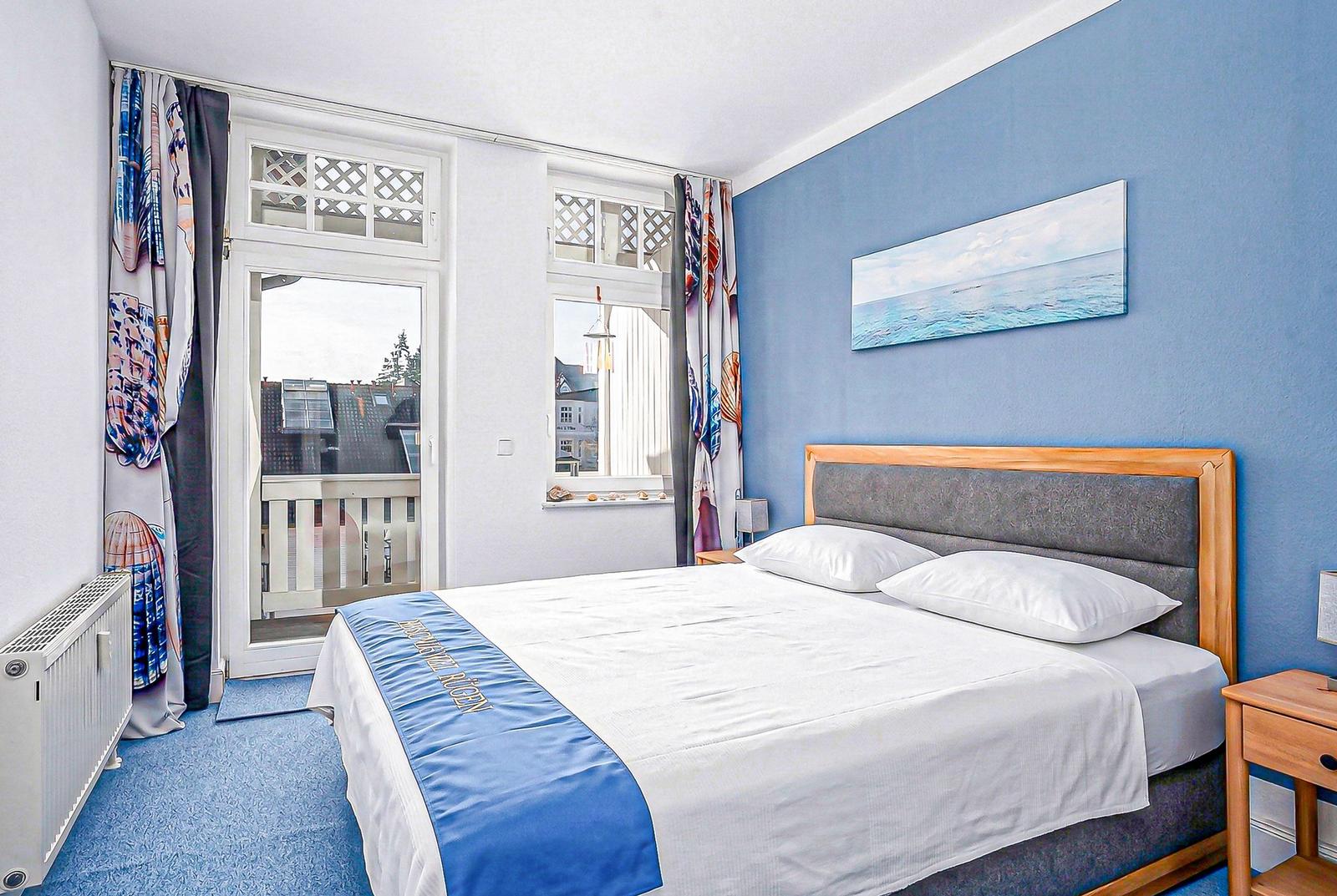 Schlafzimmer mit Bett, Fenster und Wandbild. Blauer Teppich und Vorhänge.