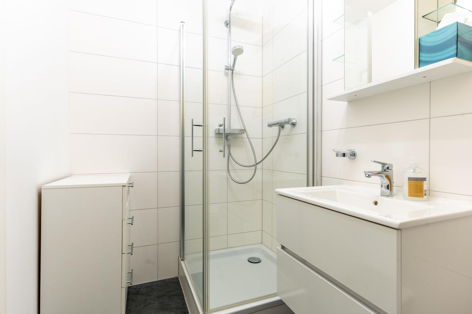 Bad mit Dusche, Waschbecken und Schrank. Fliesenwand und weiße Möbel.