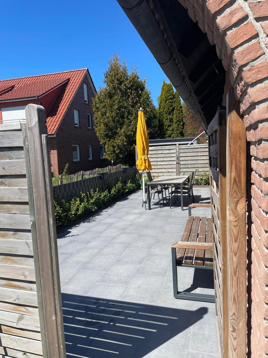 Terrasse mit Tisch, Stühlen und gelber Sonnenschirm. Holzgeländer und Ziegelmauer sichtbar.