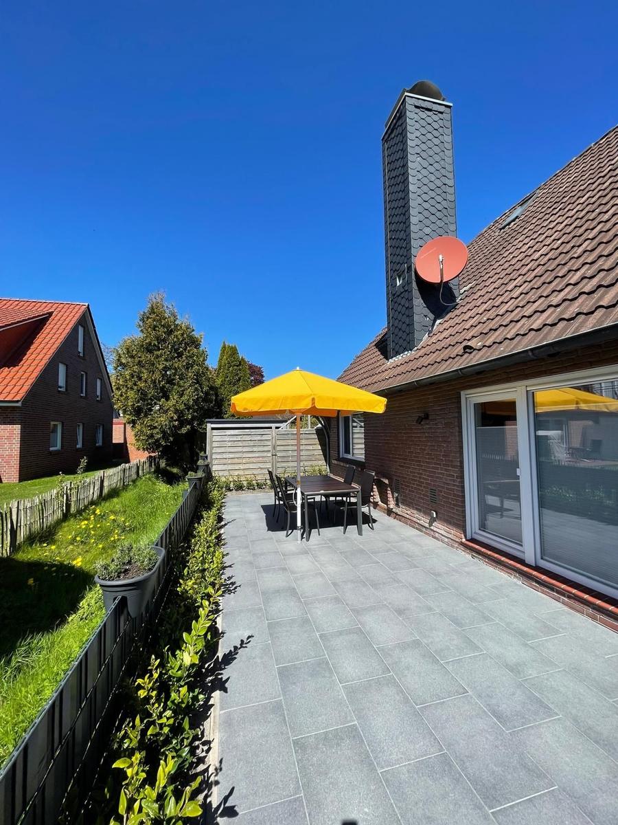 Terrasse mit gelbem Sonnenschirm und Tisch. Hintergrund: Haus mit Schornstein und Satellitenschüssel.