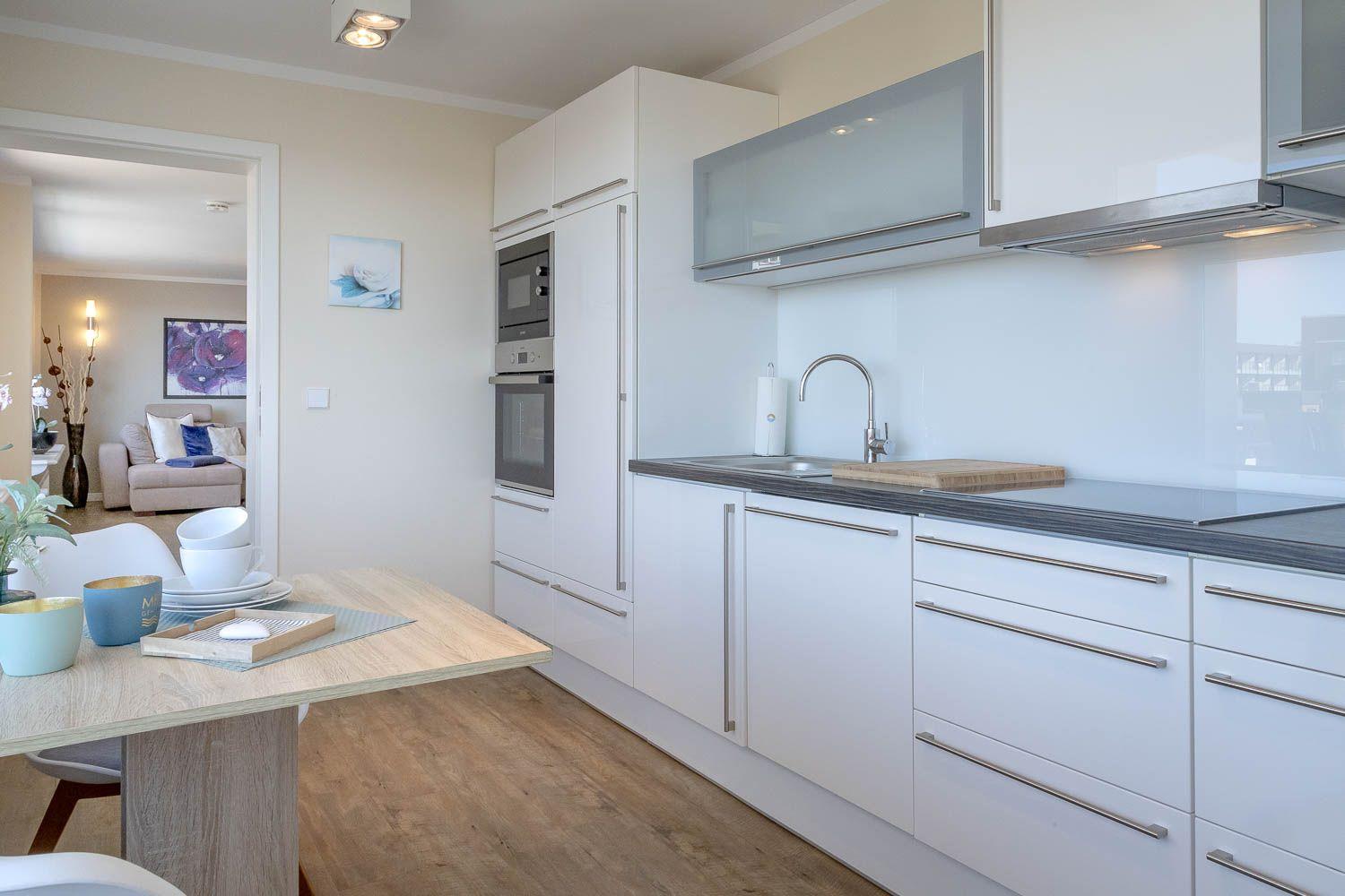 Moderne Küche mit weißen Schränken, Edelstahlarmatur und Holztisch. Wohnbereich im Hintergrund.