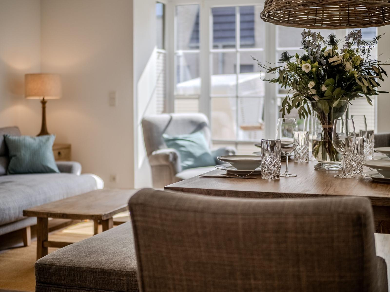 Esstisch mit Blumen, Gläsern und Stühlen. Im Hintergrund Wohnbereich mit Sofa und Sessel.