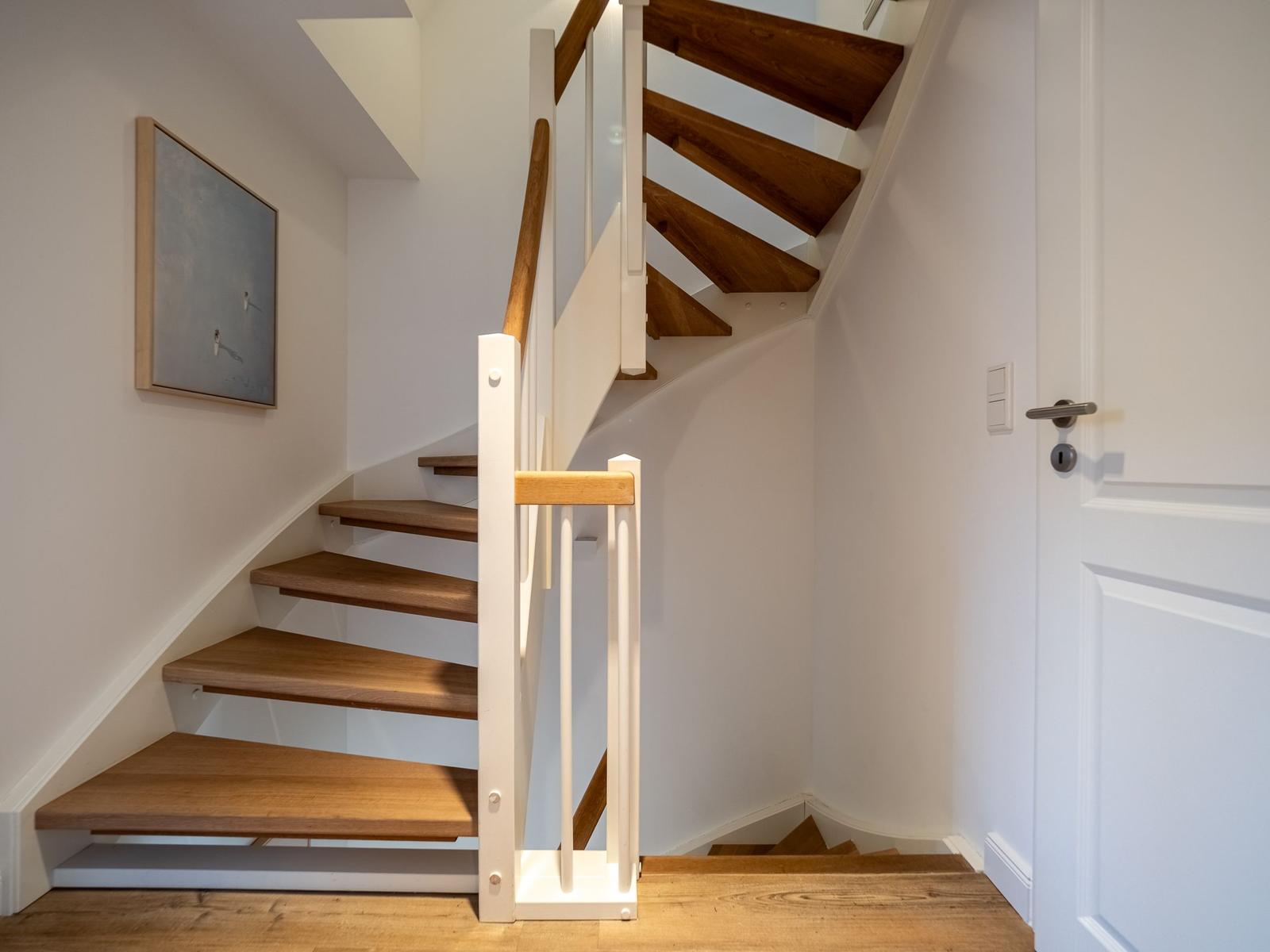 Treppenhaus mit Holztreppe und weißer Balustrade. Wandbild und Tür sind sichtbar.