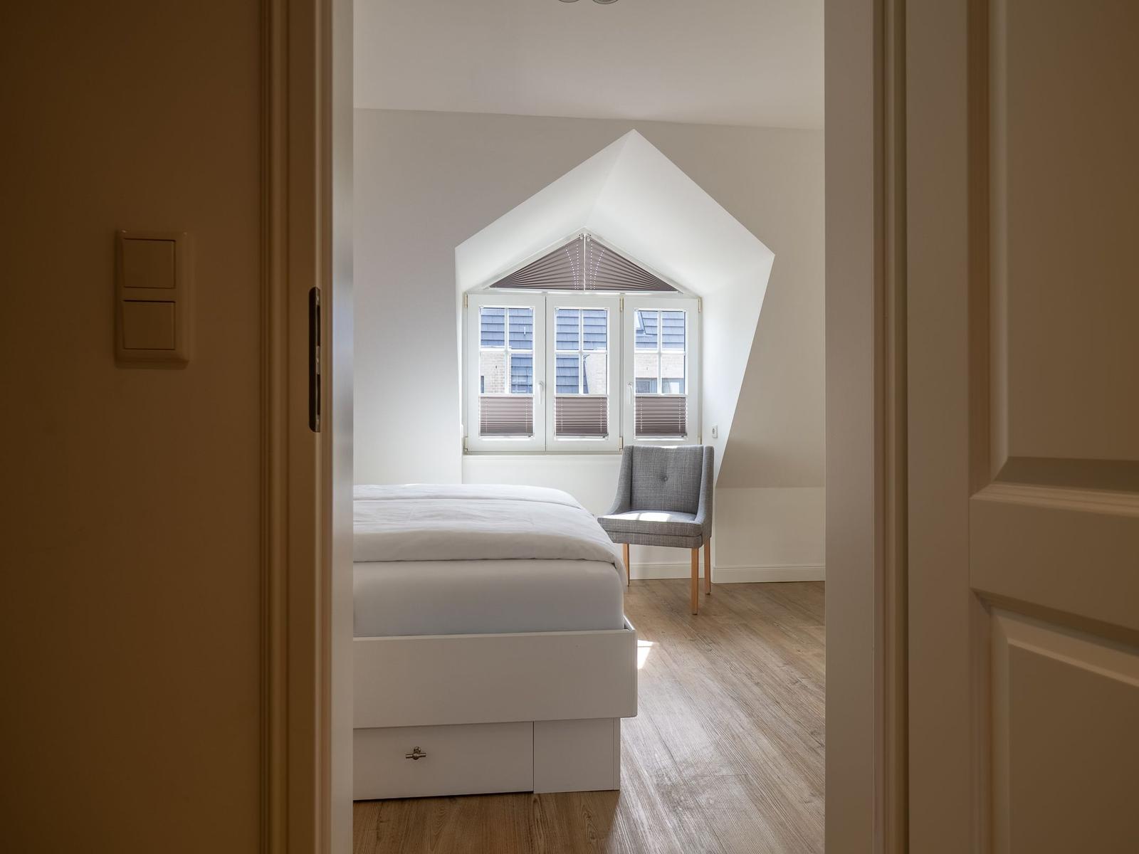 Schlafzimmer mit Dachschrägenfenster, weißem Bett und grauer Sitzgelegenheit.