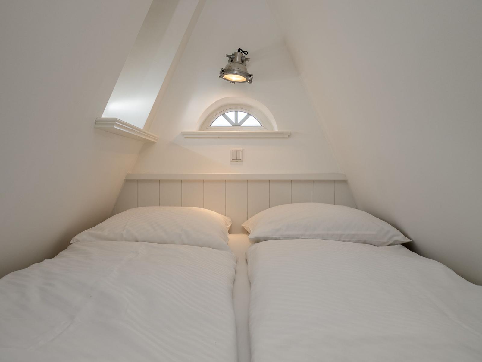 Doppelbett unter schrägen Dachschrägen mit Fenster und Deckenleuchte.