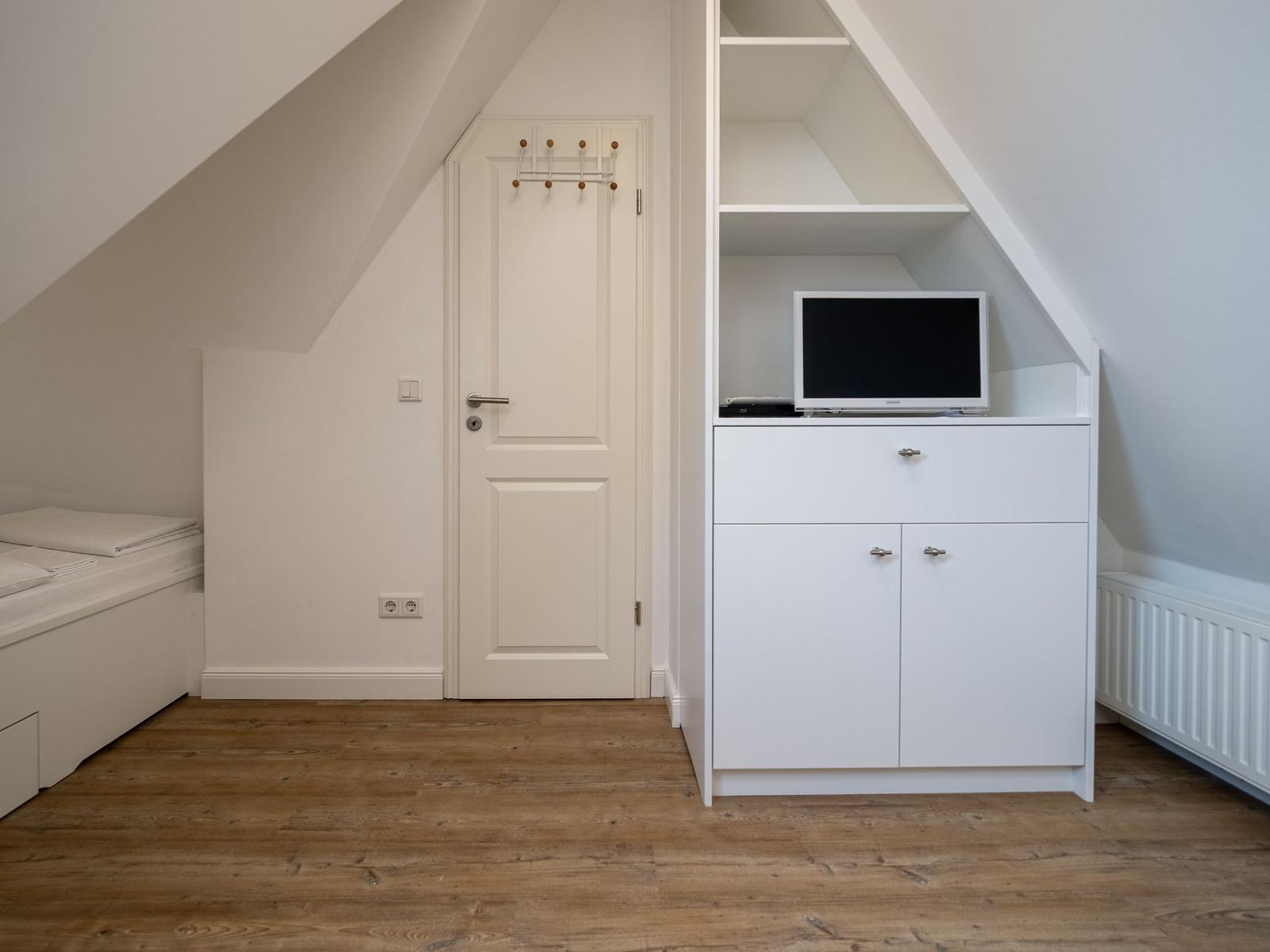 Schlafzimmer mit Dachschrägen, weißer Tür und TV-Schrank mit Schublade und Türen.