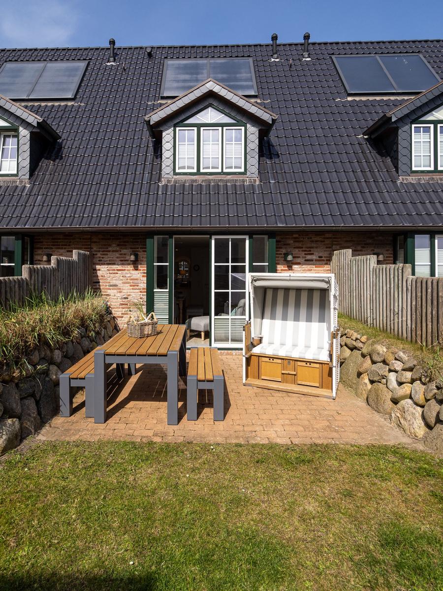 Haus mit Terrasse, Tisch, Bänken und Strandkorb. Holzwand und Dach mit Solarpanel.