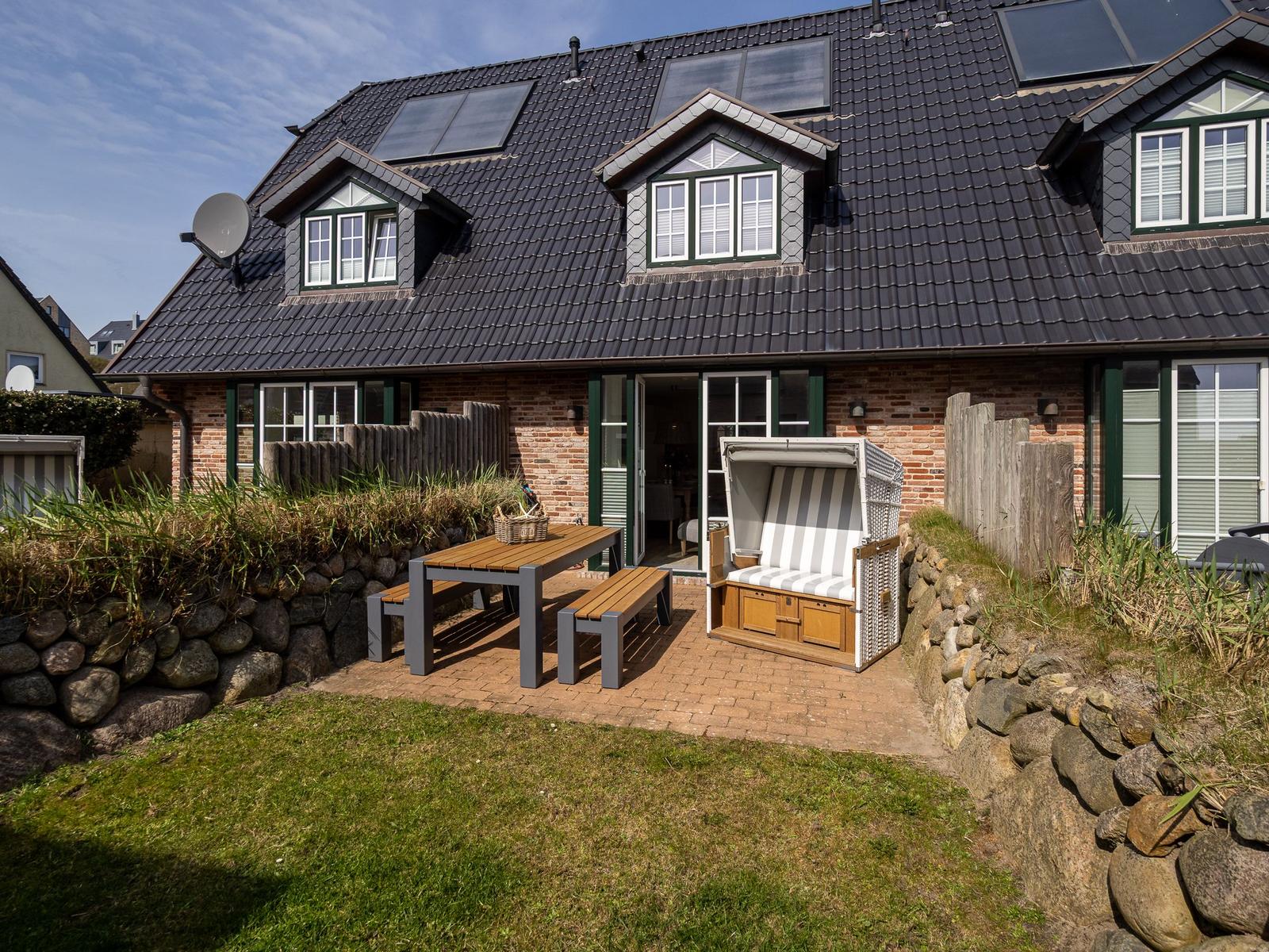 Haus mit Terrasse, Picknicktisch und Strandkorb. Schwarzes Dach, Ziegelmauer und Garten.