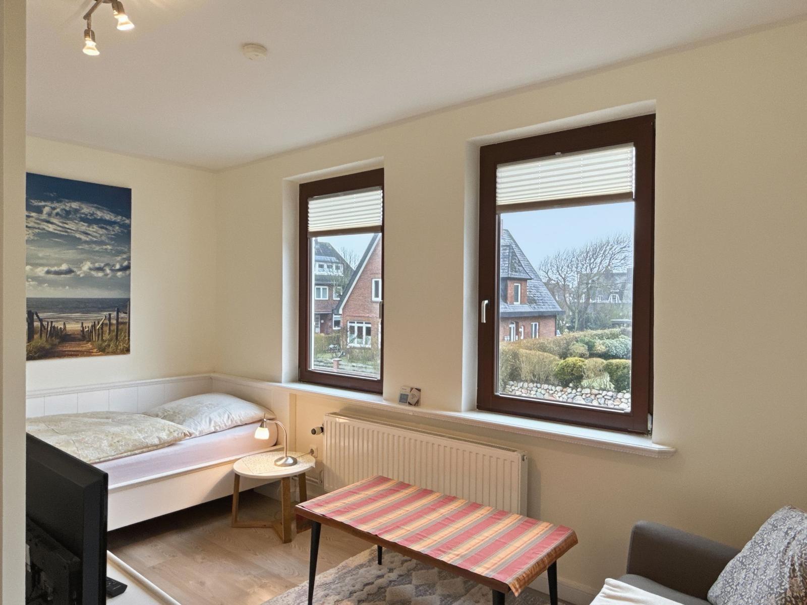 Room with bed, table, and two windows overlooking houses.