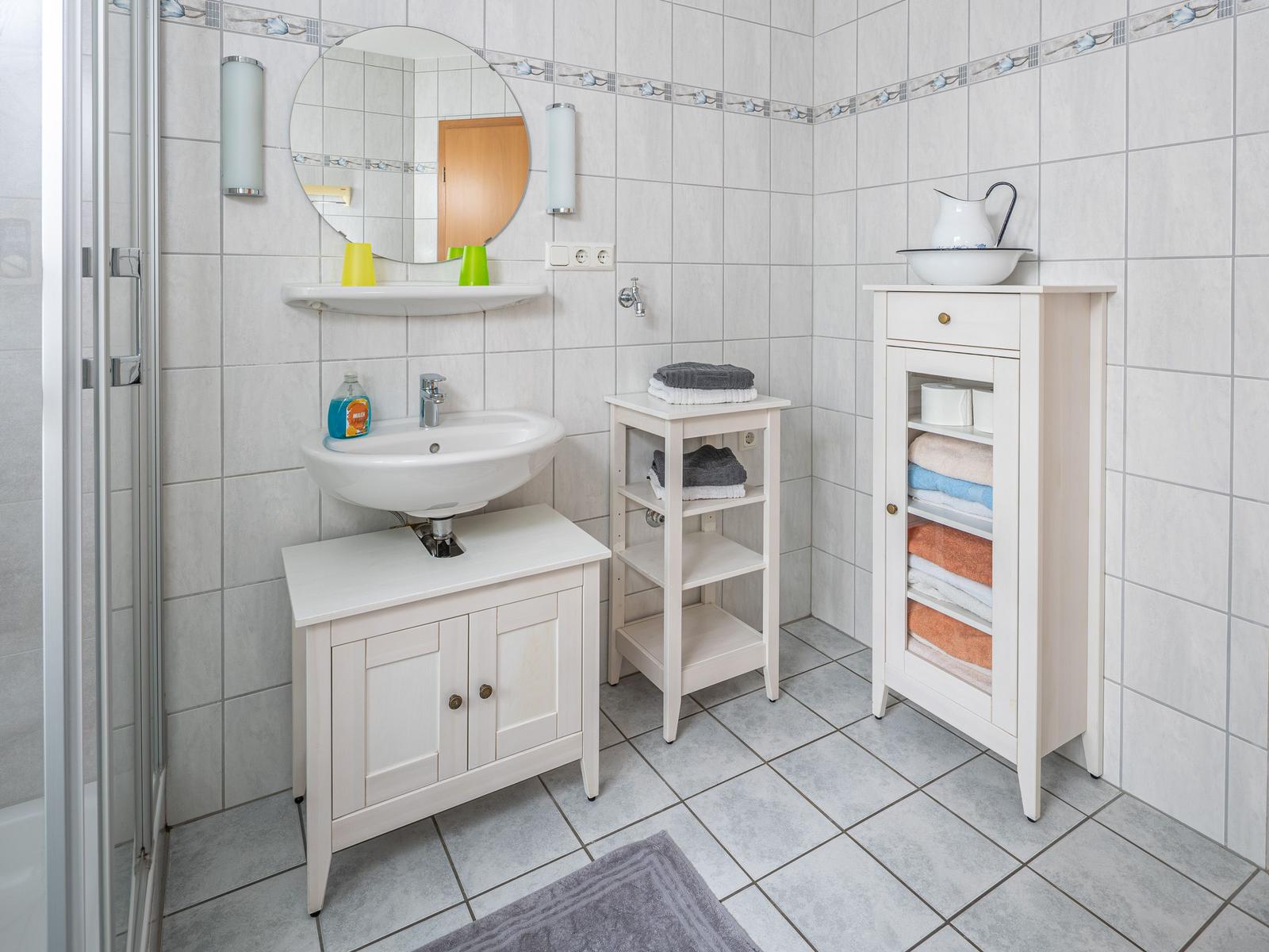 Badezimmer mit Waschbecken, Spiegel, Dusche und Handtuchregal.