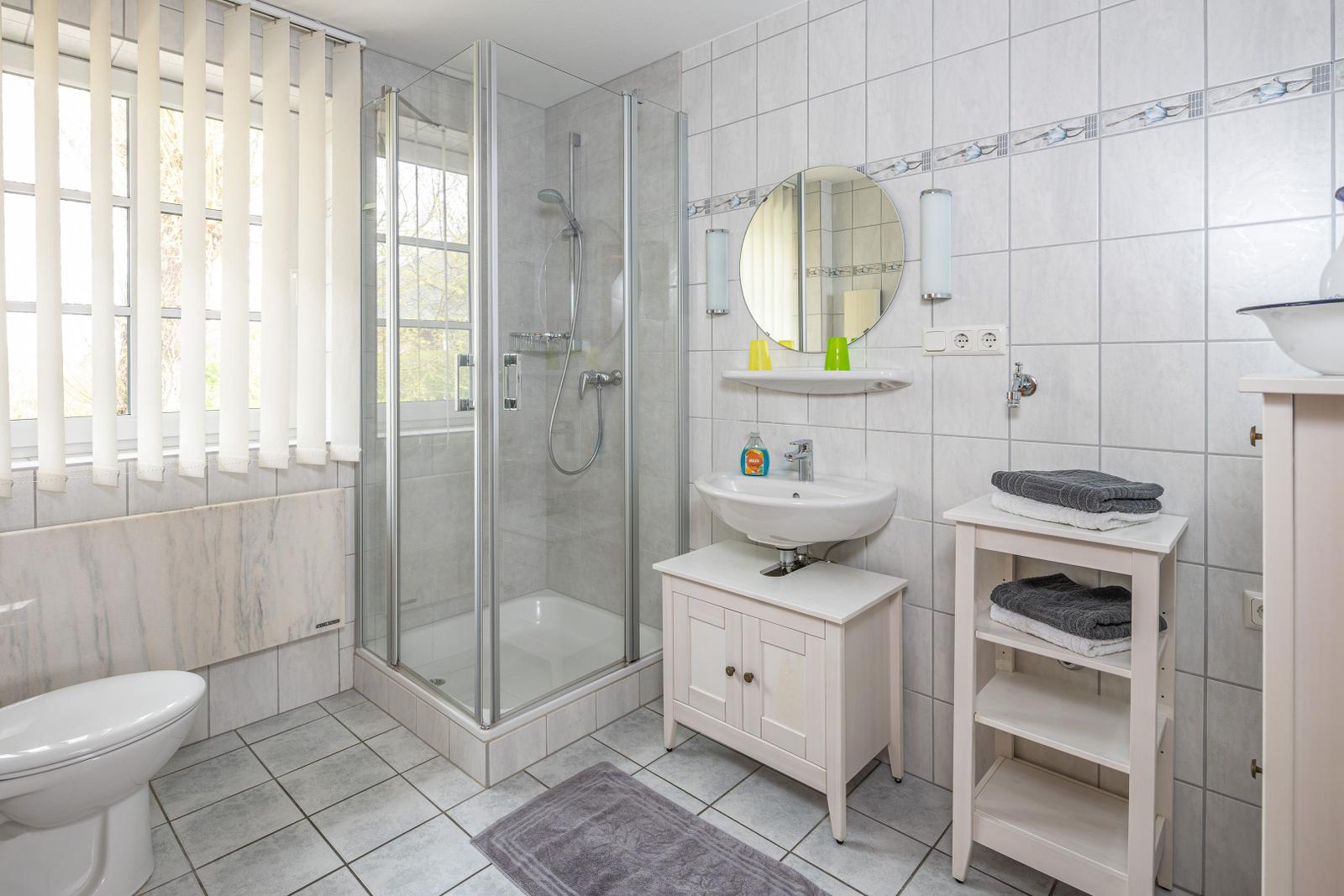Badezimmer mit Dusche, Waschbecken und Toilette. Fenster mit Jalousie.