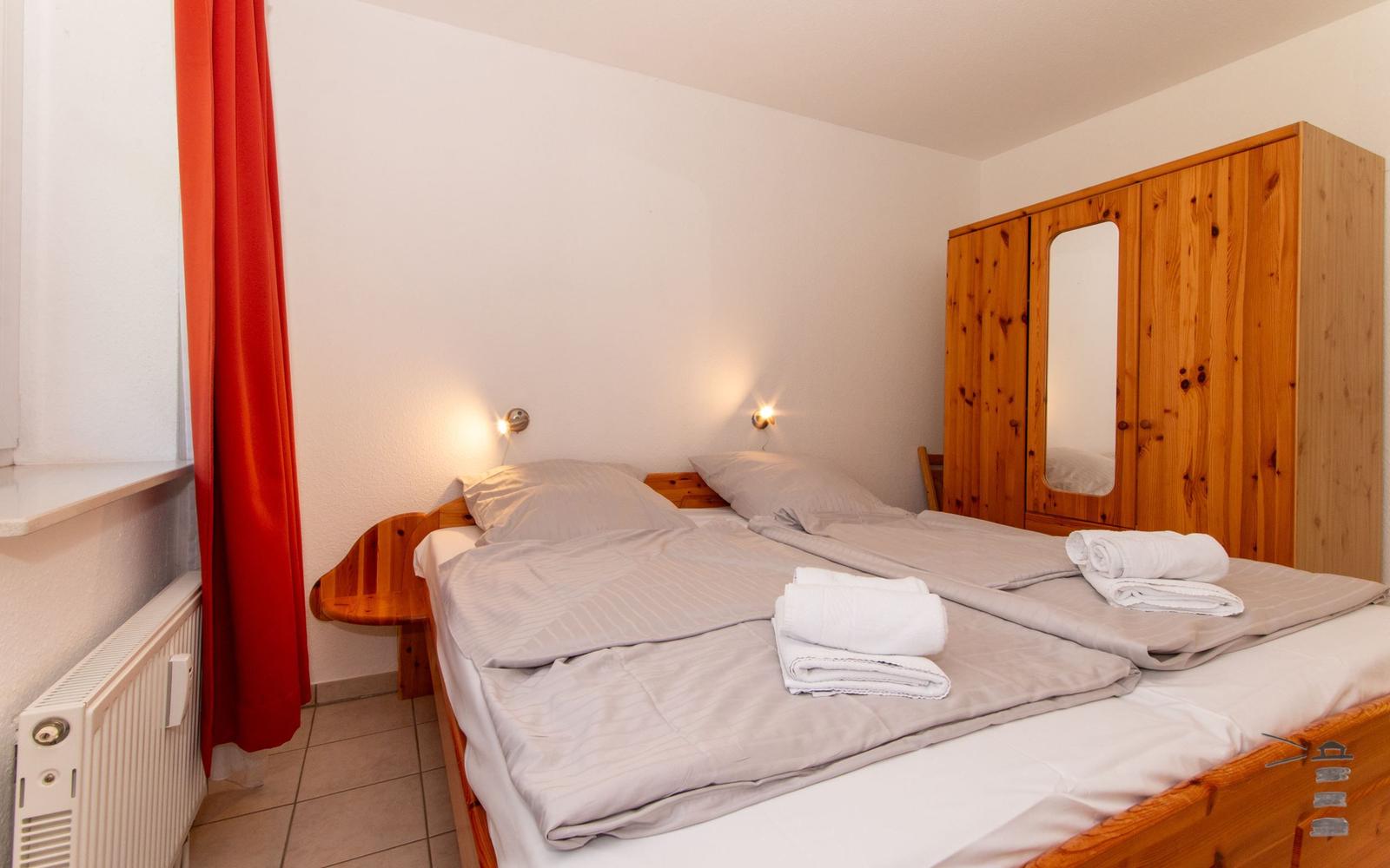 Schlafzimmer mit Doppelbett, Holzschrank und roten Vorhängen.