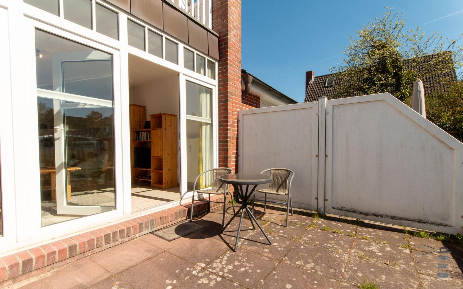 Terrasse mit Tisch und zwei Stühlen neben großem Fenster. Hinterhof mit weißer Mauer.