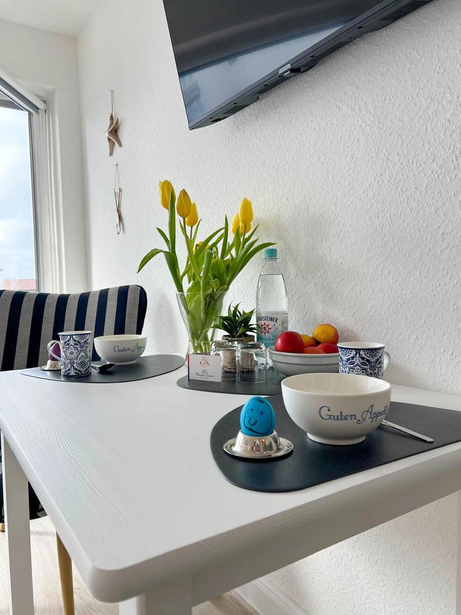 Esstisch mit Blumen, Obst und Tassen. Fernseher an der Wand.