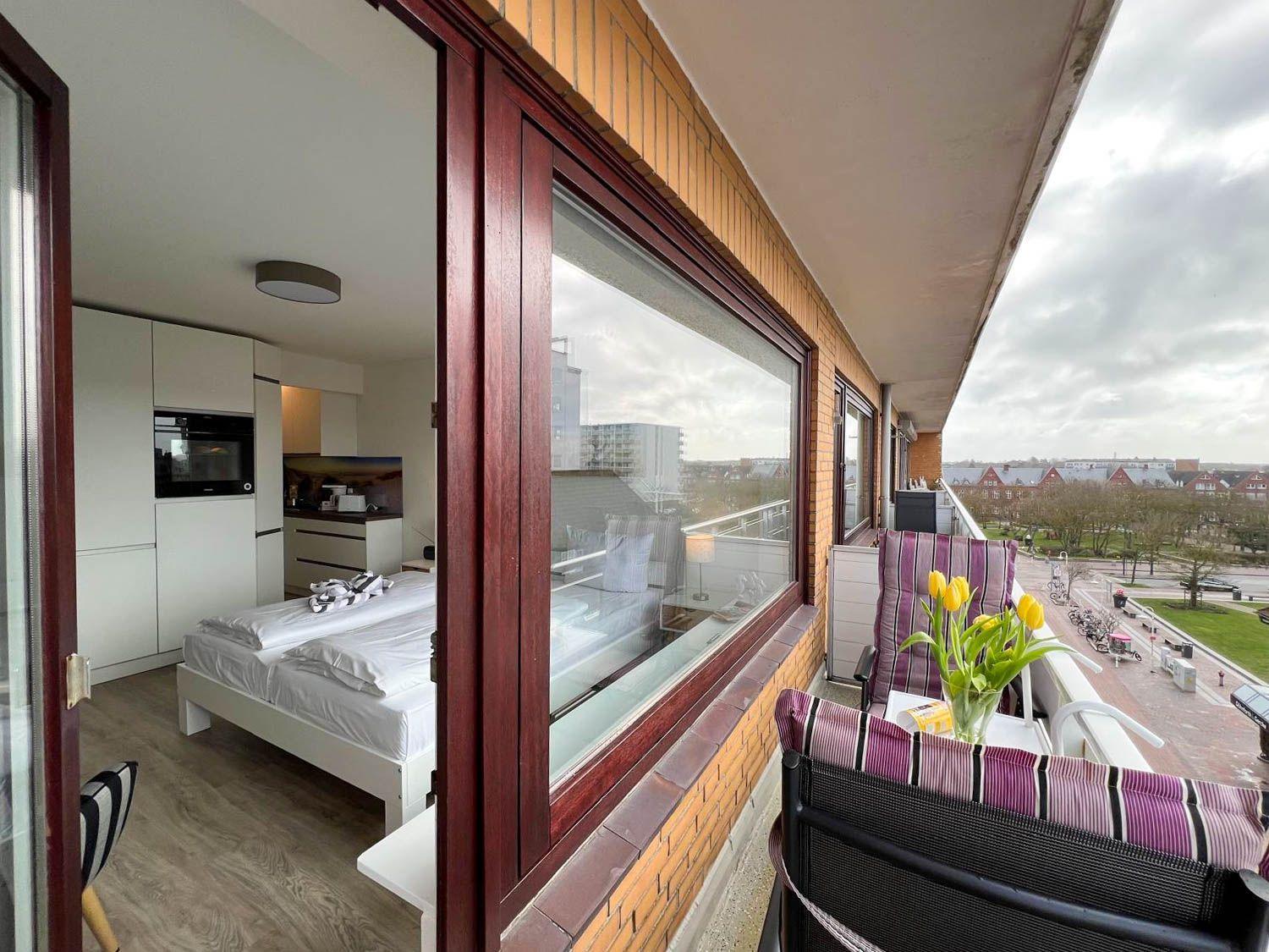 Balkon mit Stühlen und Tulpen. Blick auf Straße und Gebäude. Schlafzimmer mit Bett und Küche sichtbar.