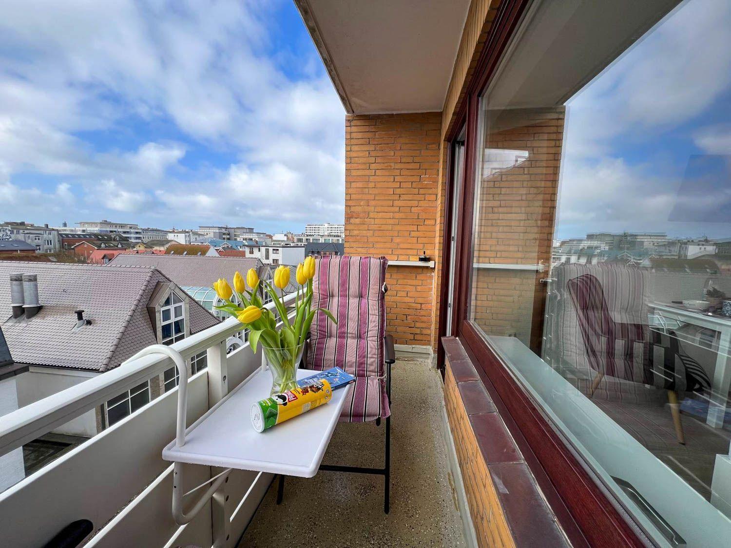 Balkon mit Stuhl, Tisch und Tulpen. Blick auf Dächer und Himmel.