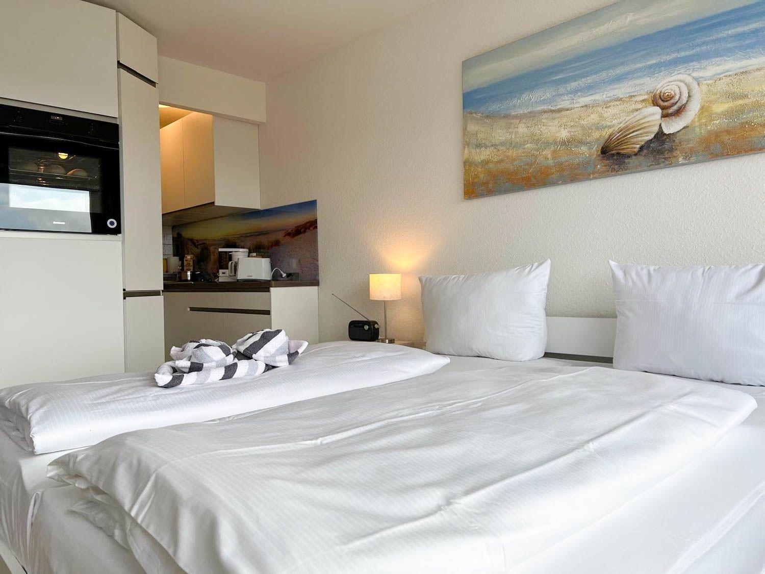 Schlafzimmer mit Bett, Nachttisch und Wandbild. Küche im Hintergrund.