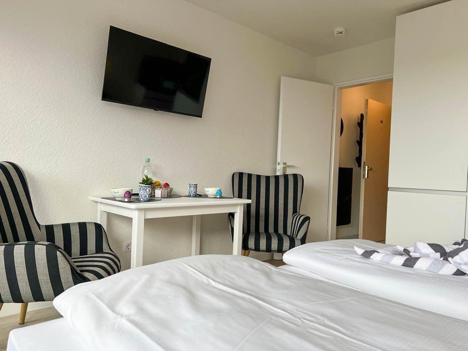 Zwei Betten, Schreibtisch mit Stühlen, Wandfernseher und Tür zum Flur.